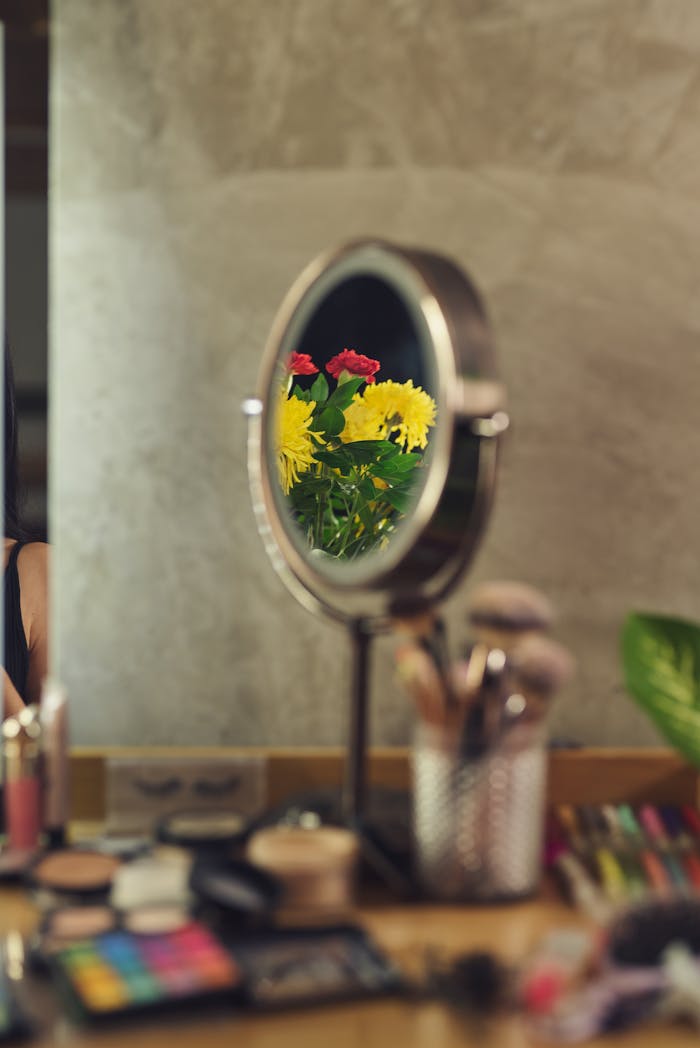 Mirror on vanity table reflects bright flowers amidst a set of cosmetics and brushes.